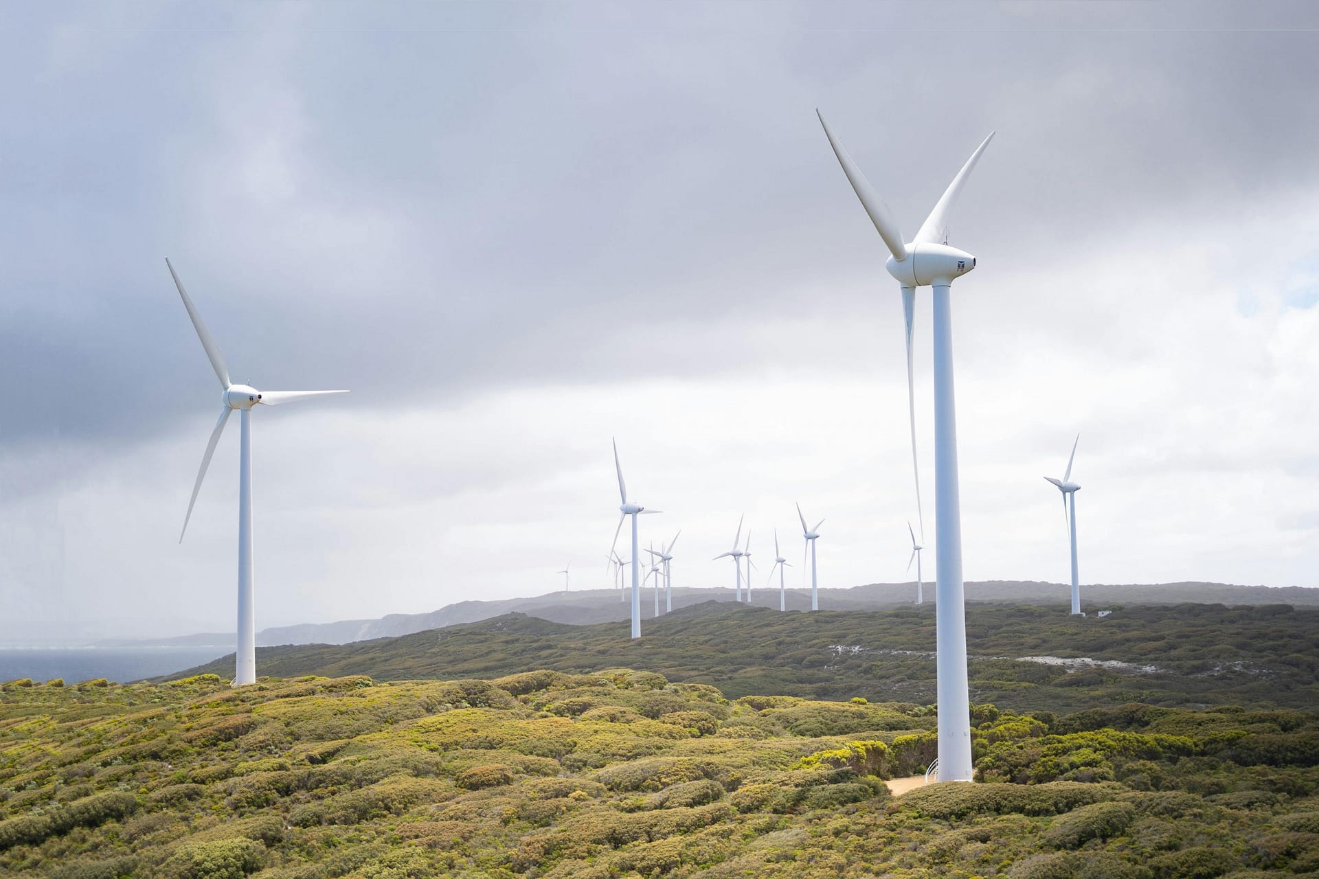 The image shows a wind farm situated on hilly, coastal terrain. Multiple large white wind turbines are positioned across the landscape it would be possible to make a digital twin of the whole coast line with accurate weather measurements all simulated in the synthetic environment of the digital twin this would provide a wealth of data for Energy Sector applications